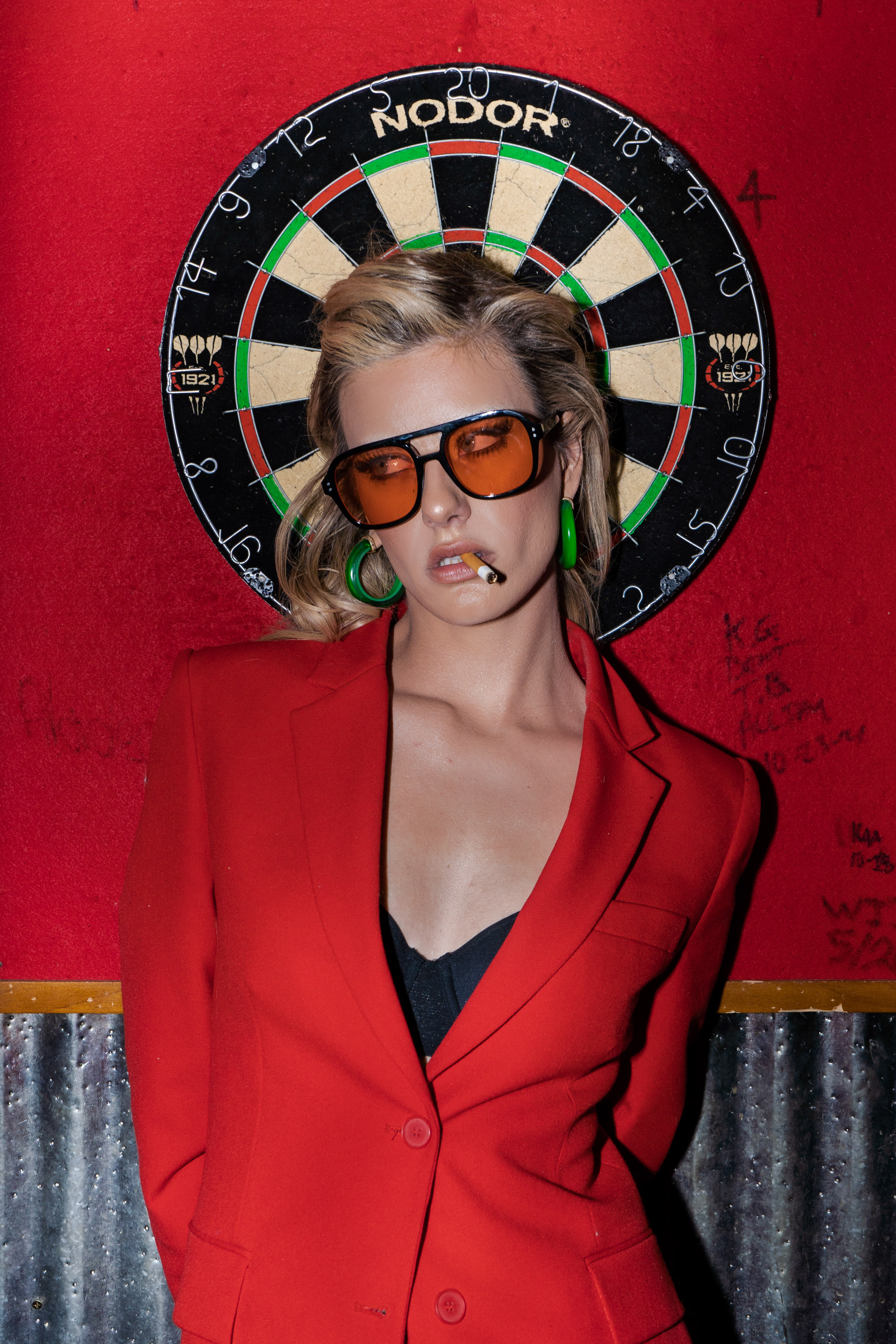 Dawn Muensterman in an oversized red blazer and amber sunglasses in front of a dartboard
